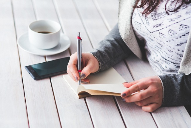 手帳に書き込む女性の手の置くにスマホやコーヒーカップが置かれている